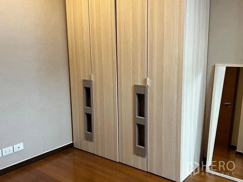 คอนโด ให้เช่า - มุมห้องนอนที่มีตู้เสื้อผ้าไม้โทนอ่อนแบบบานเปิดคู่ และกระจกแบบพกพา