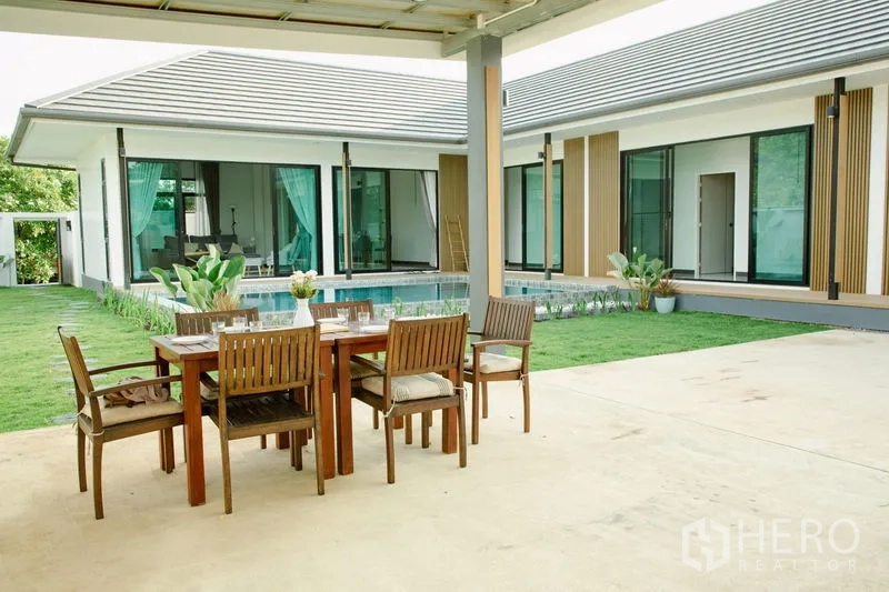 บ้าน ขายในPhra Pradaeng, Samut Prakan - เฉลียงมีหลังคาพร้อมชุดโต๊ะอาหารไม้ มองเห็นสระว่ายน้ำและสนามหญ้าของบ้านชั้นเดียวสมัยใหม่