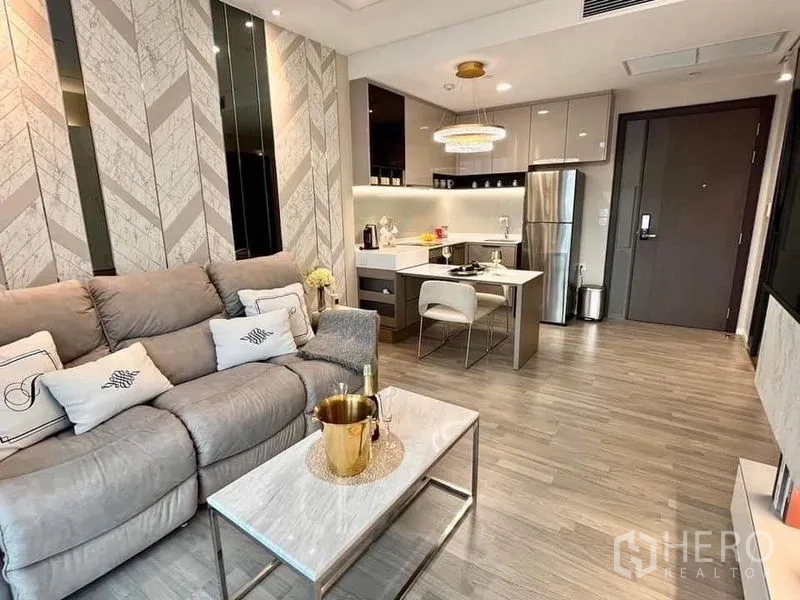 Condominium for sale in Bang Sue, Bangkok - Open-plan living room with gray sofa, marble coffee table and kitchen island with stainless fridge.