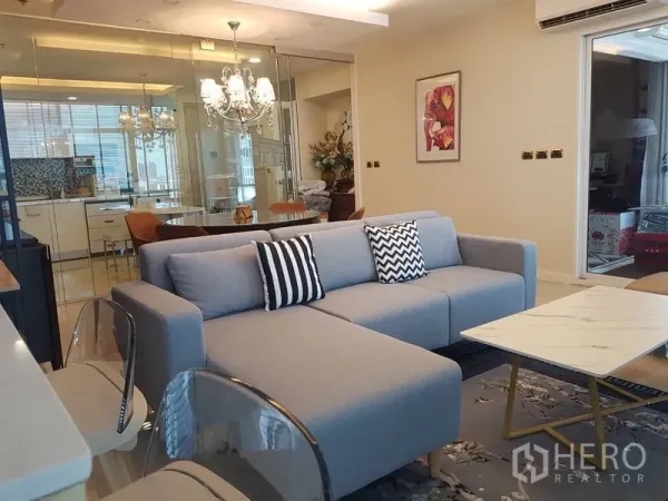 公寓 出租或出售在Bang Na, Bangkok - 采光充足的客厅，灰色转角沙发与地毯，可透过玻璃墙看到现代厨房。