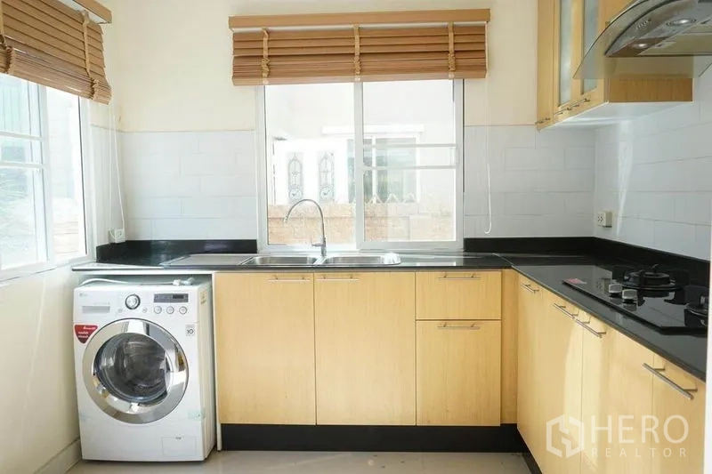 Detached House for rent in Bang Phli, Samut Prakan - Modern kitchen with light wood cabinetry, black countertops, gas hob, sink and washing machine under the window.