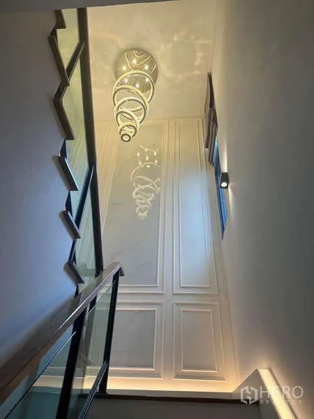 Detached House for rent in Bang Phli, Samut Prakan - Staircase with glass balustrade and statement chandelier beside a tall window.