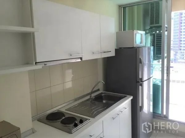 Condominium for rent in Watthana, Bangkok - Compact kitchen with white cabinets, electric hob, sink, microwave and fridge beside a balcony door.