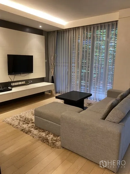 Condominium for rent in Watthana, Bangkok - Cozy living area with wall‑mounted TV, L‑shaped sofa, shag rug, and tall windows with blinds.