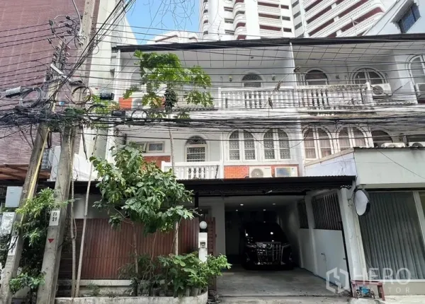 联排别墅 出租在Khlong Toei, Bangkok - 三层联排别墅立面，拱形窗与经典栏杆，带有覆盖车位的城市街景。