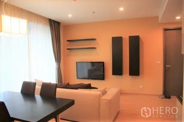 Condominium for rent in Watthana, Bangkok - Bright living room with wall-mounted TV, floating shelves, sheer floor-to-ceiling curtains and a dining table.