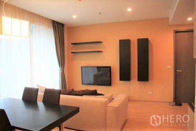 Condominium for rent in Watthana, Bangkok - Bright living room with wall-mounted TV, floating shelves, sheer floor-to-ceiling curtains and a dining table.