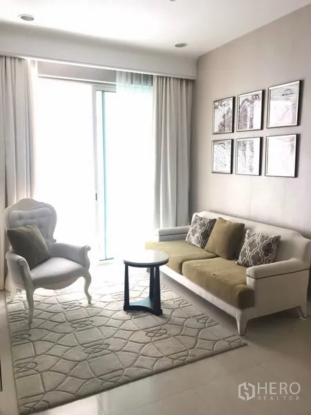 คอนโด ให้เช่าในPathum Wan, Bangkok - ห้องนั่งเล่นคอนโดสว่างไสว โซฟาสีขาว เก้าอี้วางแขน พรมลายเรขาคณิต และประตูกระจกบานเลื่อนพร้อมผ้าม่านโปร่ง