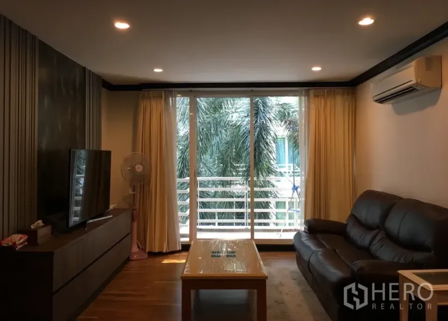 公寓 出售在Khlong Toei, Bangkok - 明亮客厅，皮沙发、电视柜，落地玻璃门通往可见棕榈树的阳台。