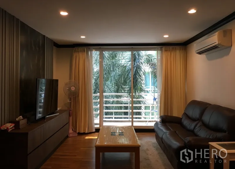 公寓 出售在Khlong Toei, Bangkok - 明亮客厅，皮沙发、电视柜，落地玻璃门通往可见棕榈树的阳台。