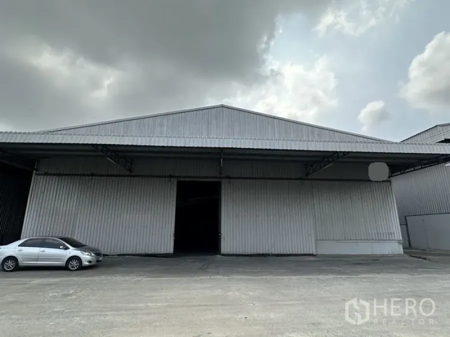 คลังสินค้า ให้เช่าในMueang Samut Prakan, Samut Prakan - ด้านหน้าโกดังผนังเหล็กพร้อมประตูบานเลื่อนกว้าง ลานคอนกรีตและรถยนต์ ใต้ท้องฟ้าครึ้ม.