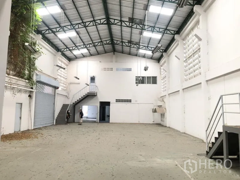 工厂 出租或出售在Bang Lamung, Chon Buri - 明亮的仓库空间，卷闸门、通往夹层的楼梯和吊灯。