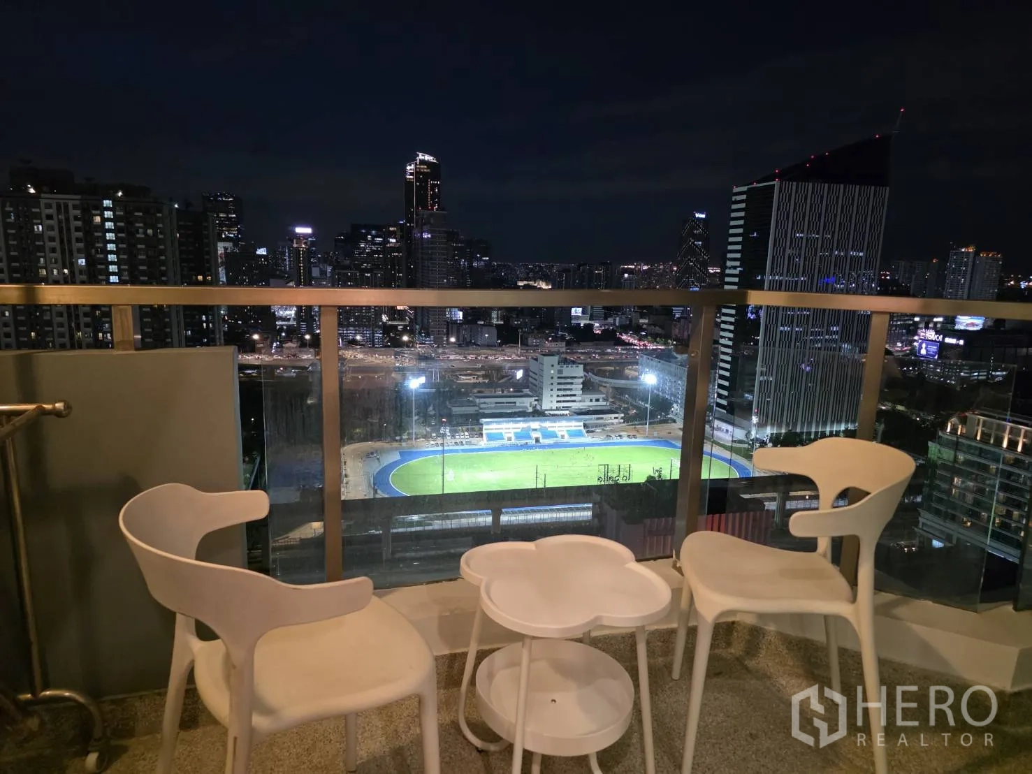 Condominium for rent in Huai Khwang, Bangkok - Night balcony with three white chairs and small tables overlooking the city lights.