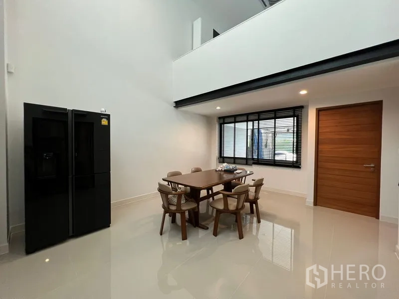 Townhouse for rent in Suan Luang, Bangkok - Bright dining area beneath a double-height ceiling with a wood door, window blinds and a black double-door fridge.