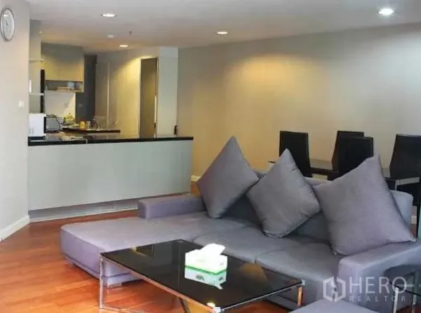 Condominium for rent in Huai Khwang, Bangkok - Open-plan living area with a grey sectional sofa, glass coffee table and wood floors beside a modern kitchen.