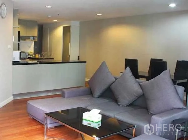 Condominium for rent in Huai Khwang, Bangkok - Open-plan living area with a grey sectional sofa, glass coffee table and wood floors beside a modern kitchen.