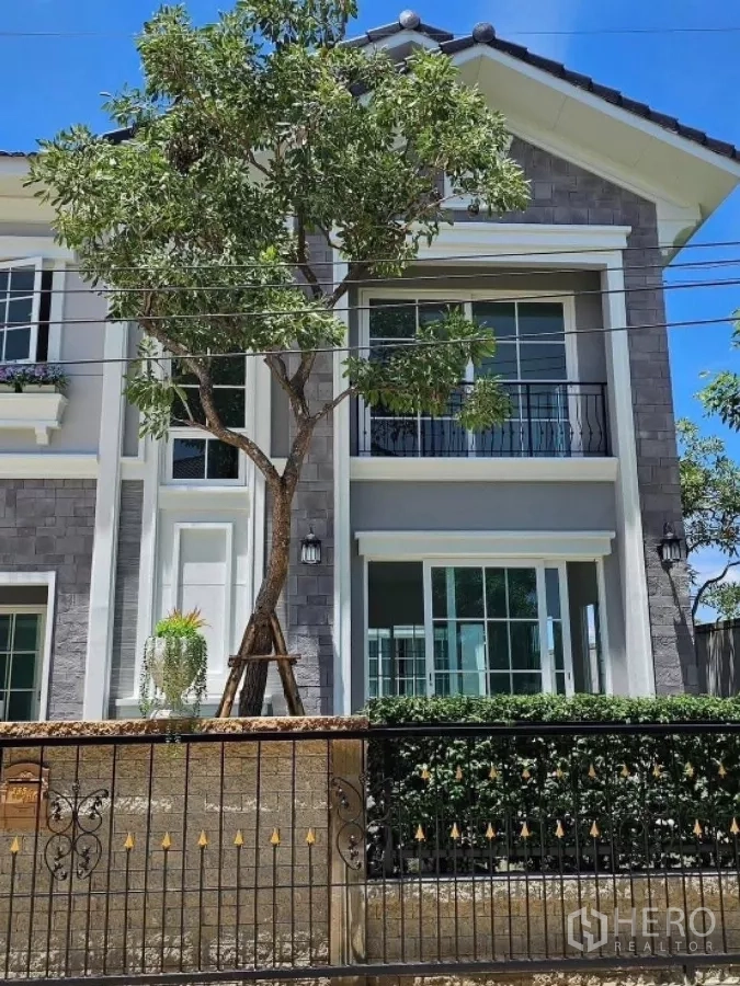 Detached House for rent in Thanyaburi, Pathum Thani - Close-up of the facade showing balcony, sliding windows and a mature tree by the front wall.