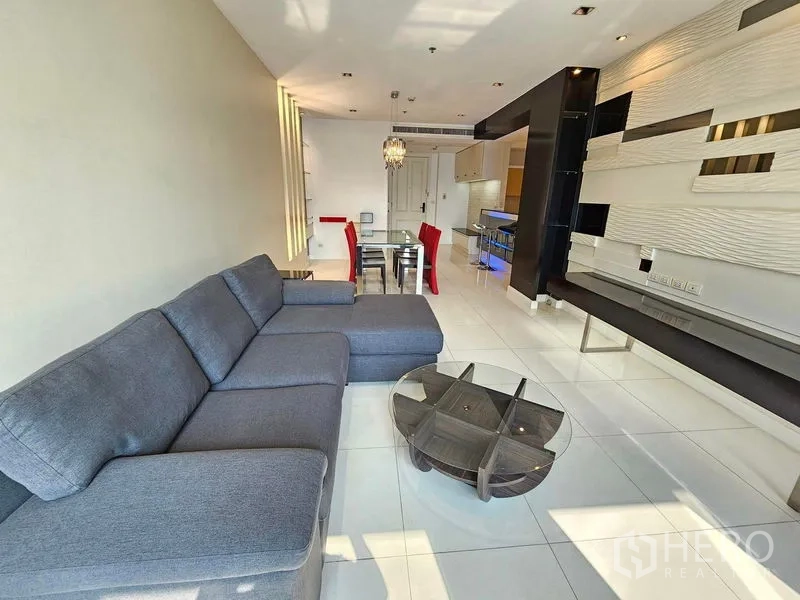 Condominium for rent or sale in Pathum Wan, Bangkok - Open-plan living and dining area with grey sectional sofa, glass coffee table and modern wall paneling.