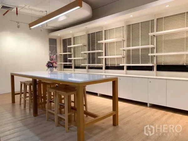 Office Space for rent in Bangkok - Collaboration zone featuring a long island table, wooden stools, wall shelving and window blinds.
