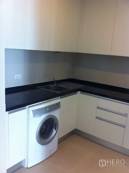 Condominium for rent or sale in Pathum Wan, Bangkok - Compact L-shaped kitchen with black countertop, white cabinets and front-loading washer.