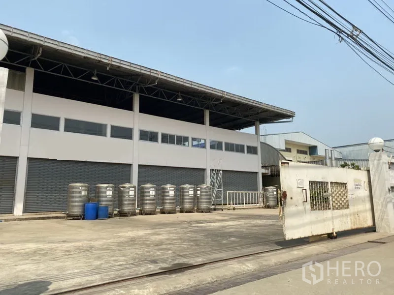 Factory for sale in Khlong Luang, Pathum Thani - Wide loading yard with roller-shutter bays at the front of a white factory building.
