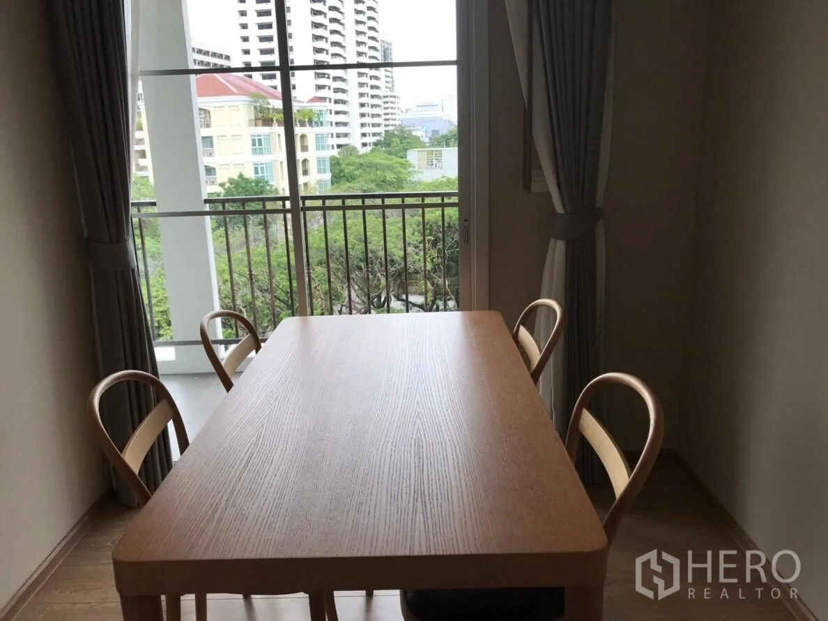 Condominium for rent in Watthana, Bangkok - Dining nook with wooden table and four chairs beside balcony window overlooking greenery.
