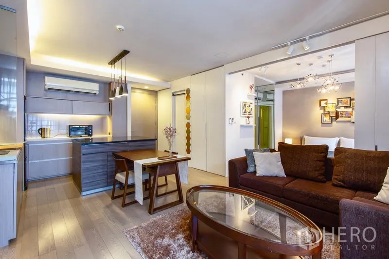 公寓 出租在Watthana, Bangkok - 开放式客厅，棕色沙发、玻璃茶几与餐桌相连，配备现代中岛厨房与吊灯。