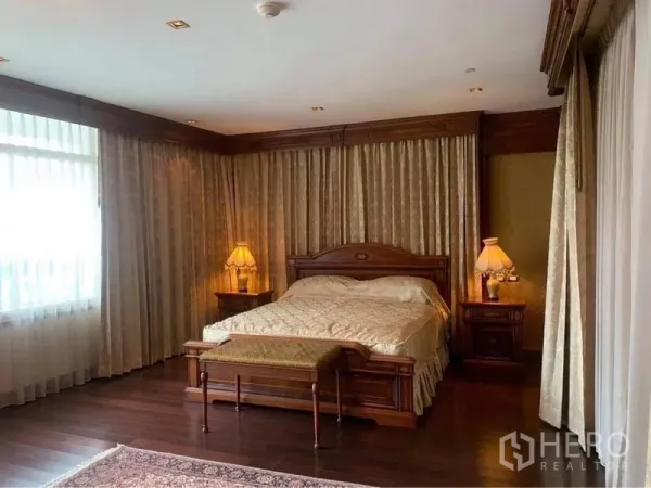 Condominium for rent in Watthana, Bangkok - Spacious master bedroom with king bed, floor-to-ceiling curtains, hardwood flooring, bedside lamps, and a bench at the foot of the bed.