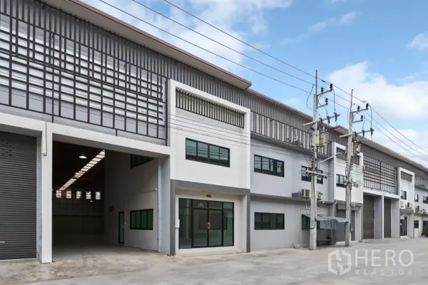 โรงงาน ให้เช่าในMueang Samut Sakhon, Samut Sakhon - แถวอาคารโรงงานหลายยูนิต มีประตูม้วนสูงและถนนภายในกว้าง หนึ่งยูนิตเปิดอยู่.