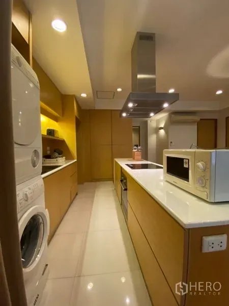 公寓 出租在Watthana, Bangkok - 走廊式厨房，白色岛台、炉灶与烟机、嵌入式柜体及叠放洗烘机。