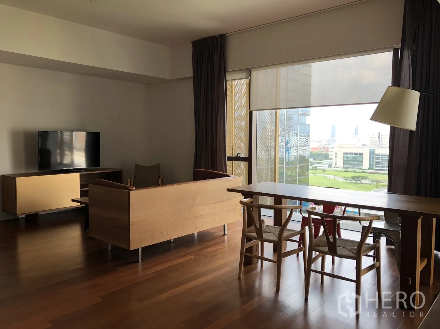 公寓 出租在Pathum Wan, Bangkok - 开放式客餐厅，实木地板、电视柜，落地窗俯瞰城市与绿地景观