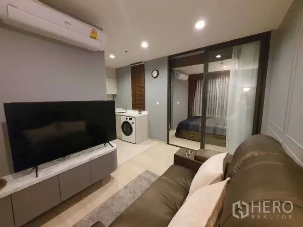 Condominium for rent - Modern living room at Life One Wireless with sofa, TV, kitchenette with washer, and glass door to the bedroom.