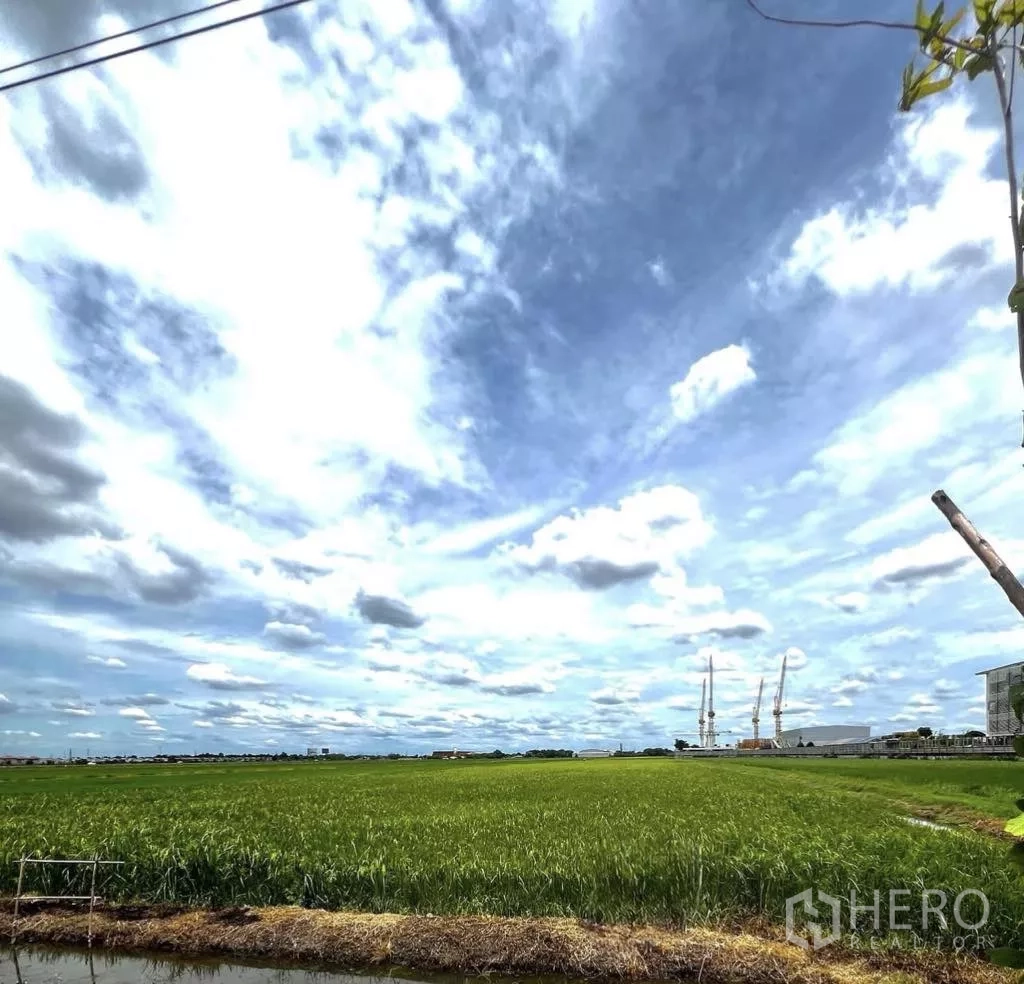Land for sale in Lam Luk Ka, Pathum Thani - Expansive green field in Lam Luk Ka with a drainage canal in the foreground and distant industrial cranes under a wide cloudy sky.