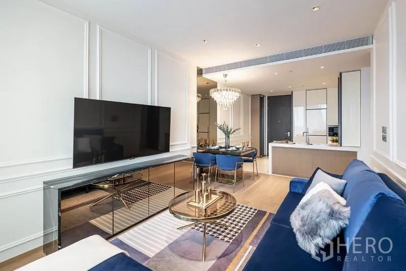 คอนโด ให้เช่าในPathum Wan, Bangkok - พื้นที่นั่งเล่นพร้อมตู้วางทีวีขนาดใหญ่ โซฟาสีน้ำเงิน และครัวแบบเปิดด้านหลัง