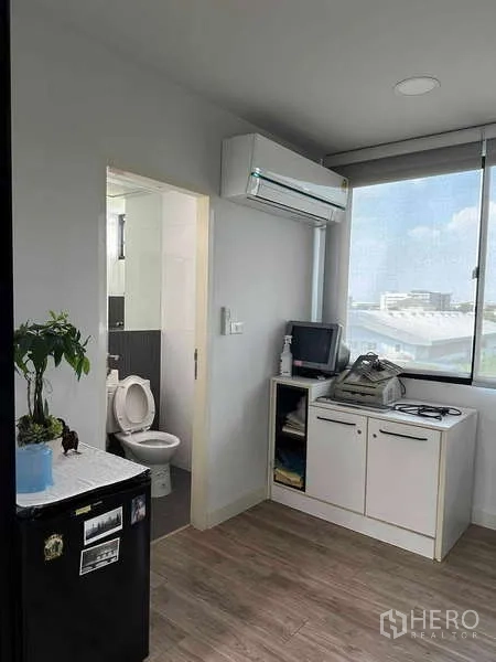 Home Office for rent in Bueng Kum, Bangkok - Small kitchenette beside a restroom, wall-mounted AC, and wide window providing daylight.