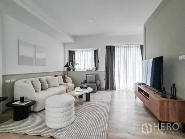 联排别墅 出租在Prawet, Bangkok - 采光充足的客厅，弧形沙发、电视柜、条纹地毯与薄纱窗帘。