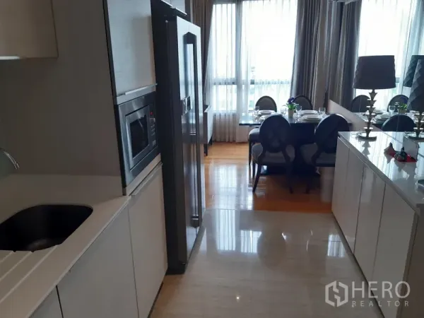 Condominium for rent in Watthana, Bangkok - Galley kitchen with white cabinetry, built-in microwave and tall fridge leading to a bright dining area with large windows.