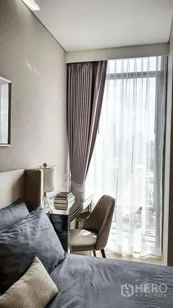 Condominium for sale in Khlong Toei, Bangkok - Bedroom corner with study desk and chair beside floor-to-ceiling curtained window letting in city light.