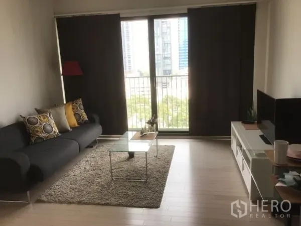 Condominium for rent or sale in Watthana, Bangkok - Bright living room with dark sofa, glass coffee table, rug, and floor-to-ceiling balcony doors with a city view.