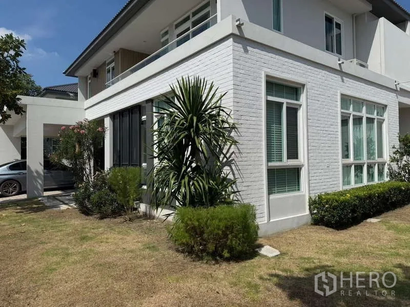 Detached House for rent or sale in Bang Bua Thong, Nonthaburi - Side exterior with white brick cladding, tall windows, and a lawn with tropical plants.
