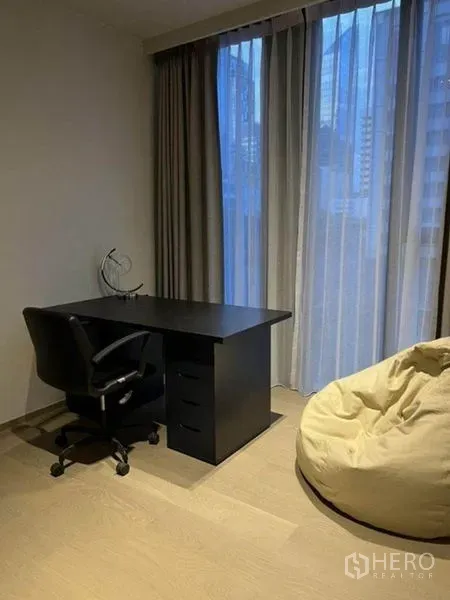 Condominium for rent in Pathum Wan, Bangkok - Work-from-home corner with black desk, chair and beanbag beside floor-to-ceiling curtains.