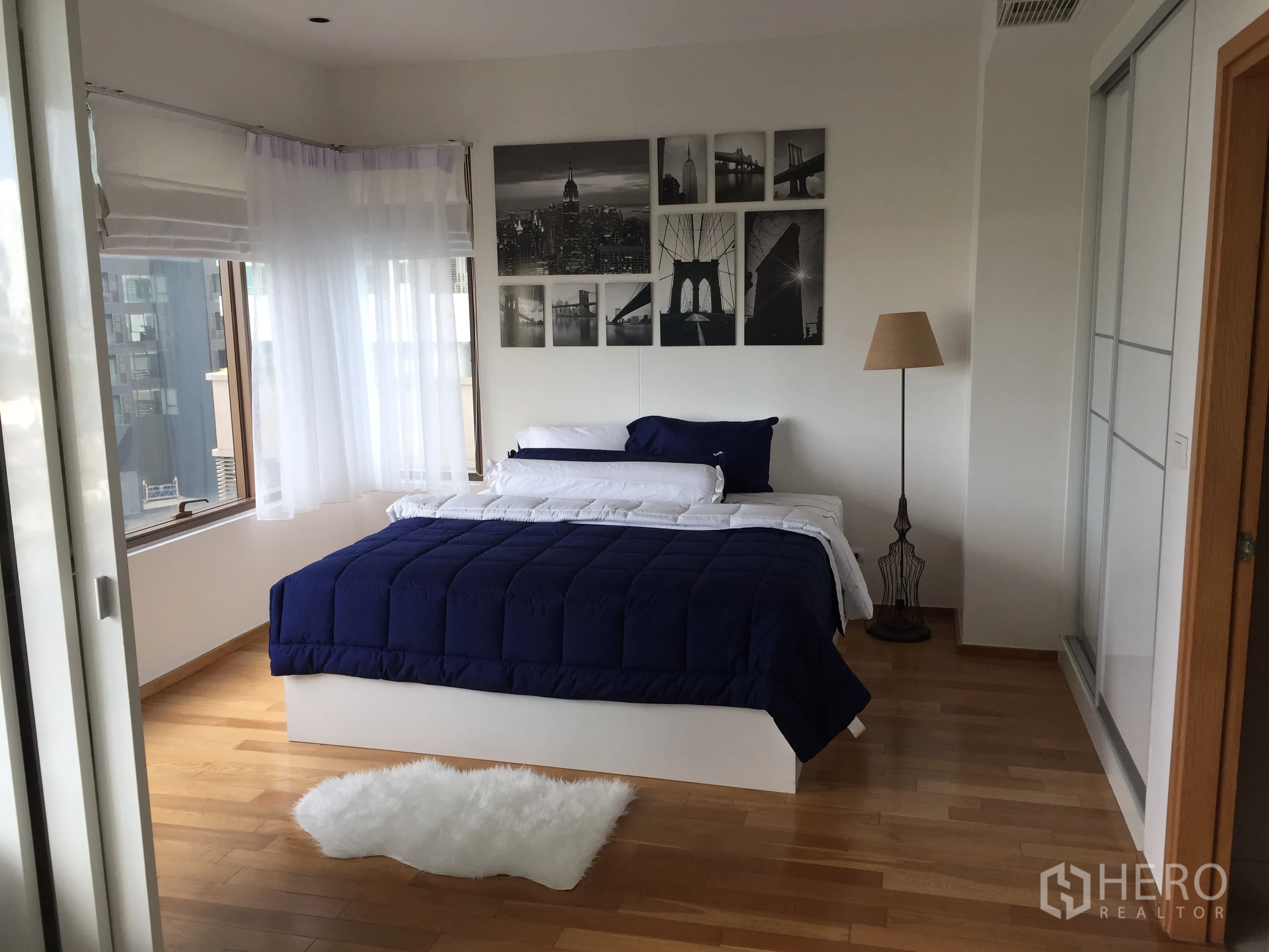 คอนโด ให้เช่าในKhlong Toei, Bangkok - ห้องนอนสว่างด้วยหน้าต่างมุม เตียงผ้าห่มสีน้ำเงิน ม่านโปร่ง พื้นไม้ และภาพตกแต่งผนัง