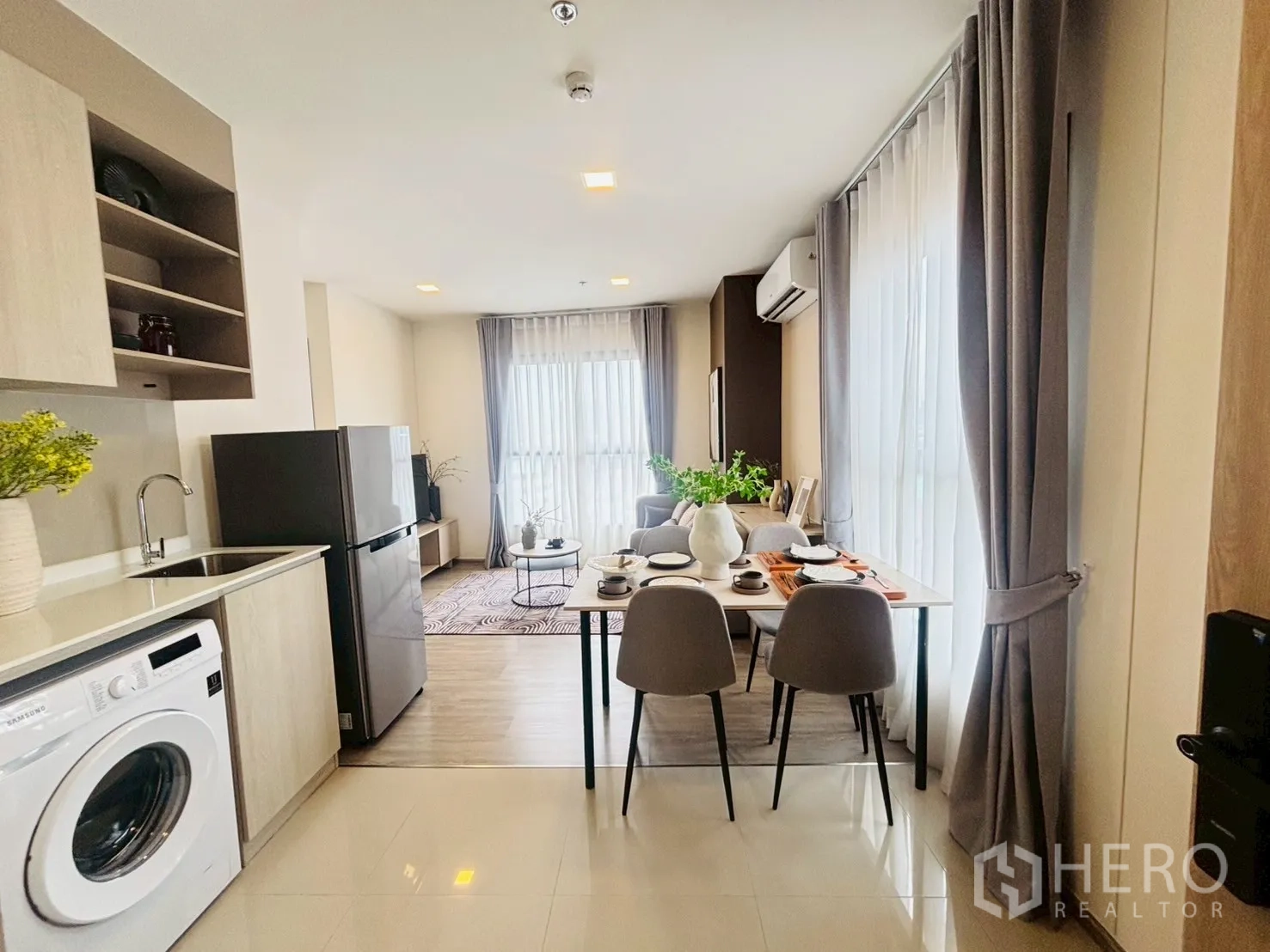 Condominium for rent in Watthana, Bangkok - Open-plan kitchen with washing machine, fridge, and a dining table beside floor-to-ceiling windows leading to a cozy living area.