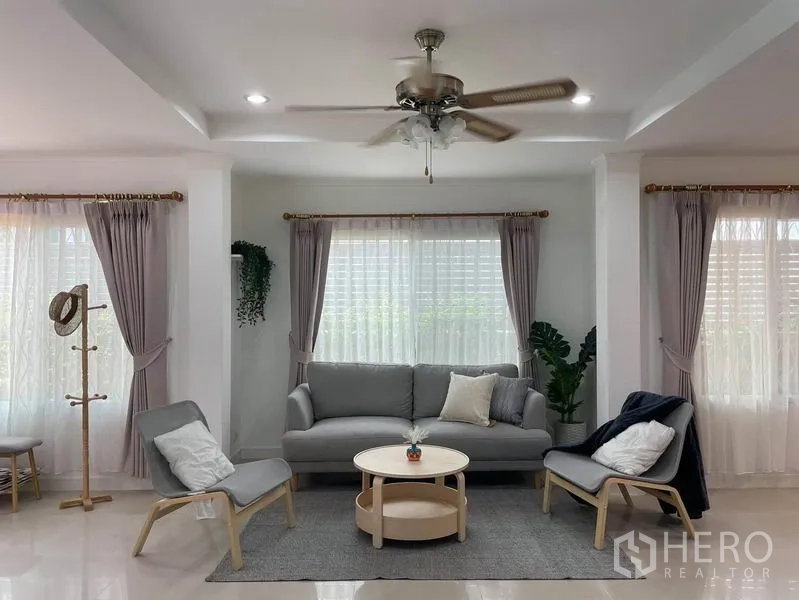บ้าน ให้เช่าในSuan Luang, Bangkok - ห้องนั่งเล่นสว่างเพดานติดพัดลม ชุดโซฟาสีเทา เก้าอี้พักผ่อน และหน้าต่างผ้าม่านบานใหญ่.