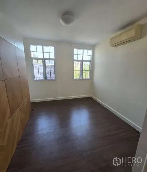 Townhouse for sale in Watthana, Bangkok - Cozy bedroom with twin windows, wood floors and a wall-mounted air conditioner.