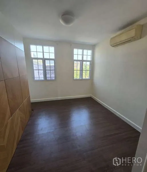 Townhouse for sale in Watthana, Bangkok - Cozy bedroom with twin windows, wood floors and a wall-mounted air conditioner.