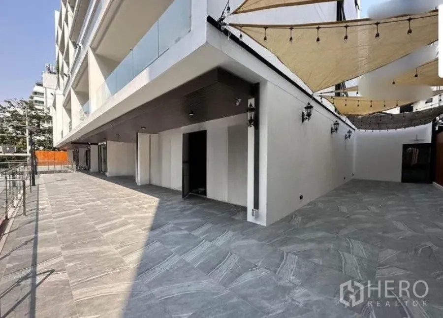 พื้นที่ขายปลีก ให้เช่า - เทอเรสชั้นล่างกว้าง ปูกระเบื้องสีเทา มีผ้าใบกันแดดและหน้าร้านกว้างใต้ตัวอาคารสมัยใหม่