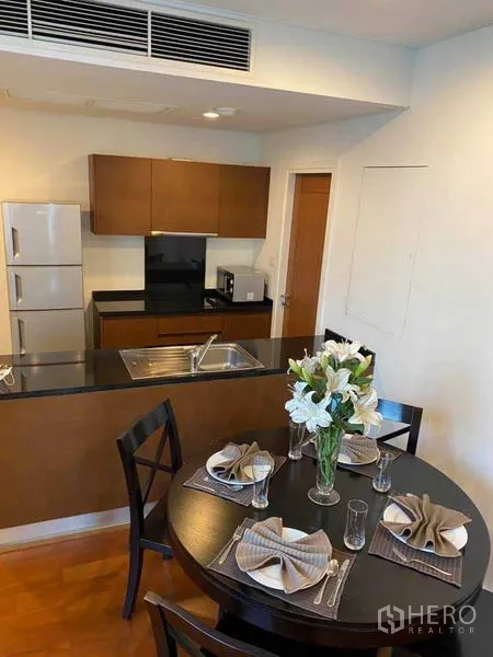 Condominium for rent in Watthana, Bangkok - Open kitchen with black countertop, sink, fridge, and a four-seat round dining table with flowers.
