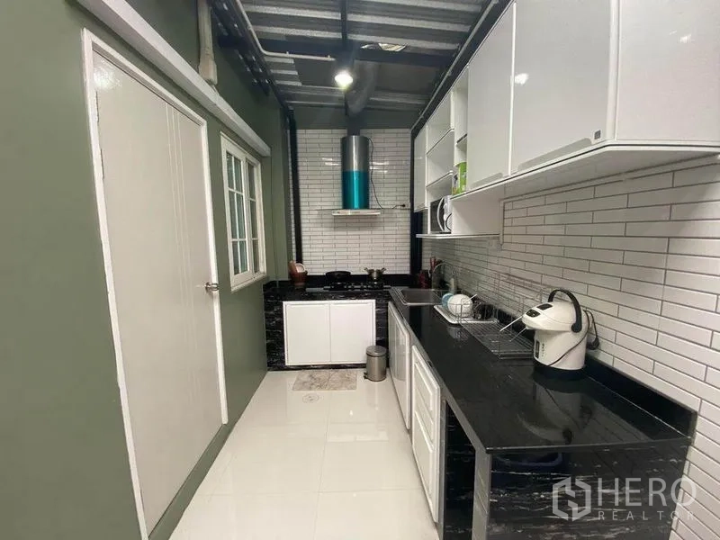 Townhouse for sale in Saphan Sung, Bangkok - Modern galley kitchen with white cabinetry, black stone countertops, subway tile backsplash and a stainless hood.