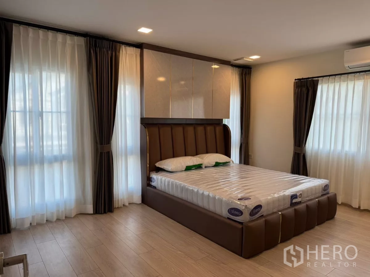 Detached House for rent in Bang Phli, Samut Prakan - Spacious master bedroom with an upholstered bed, large windows with sheer and blackout curtains, and wood-look flooring.